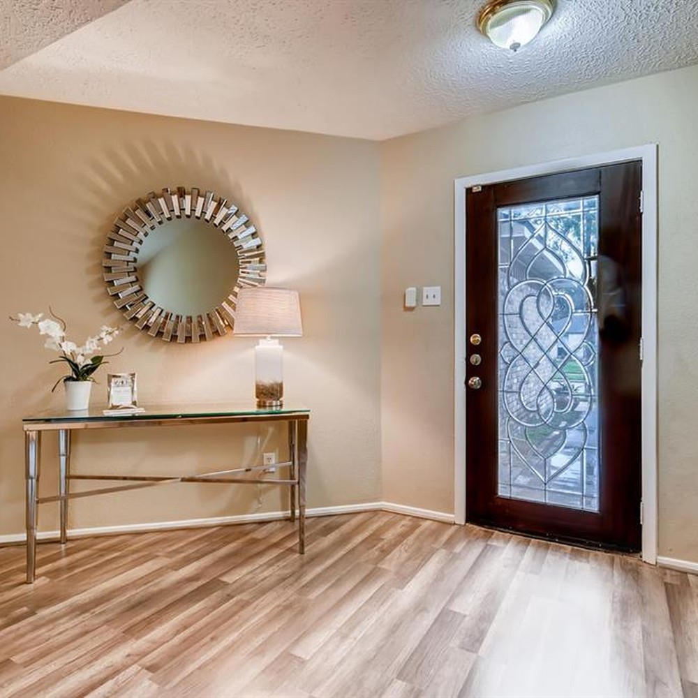 Foyer with glass entry table. Silver mirror hung by door. Lamp and white flowers on table.