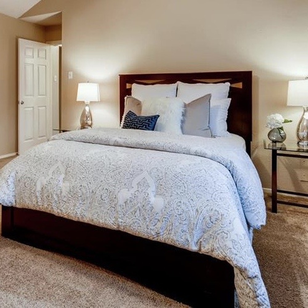 Primary bedroom with black wood bed frame. White and gray comforter, and blue and silver pillows. Glass nightstands with lamps.