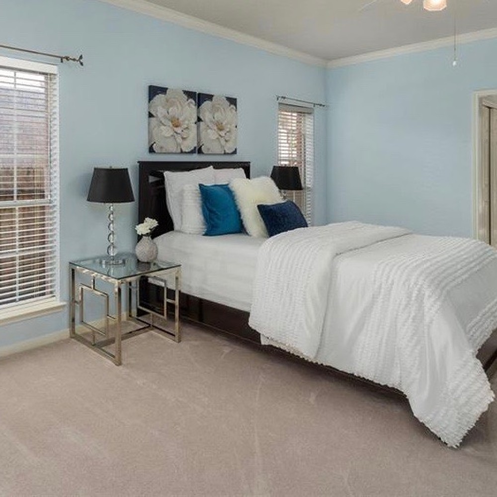 Black bed frame with white comforter. Blue and white pillows on bed. Glass nightstands with silver base.