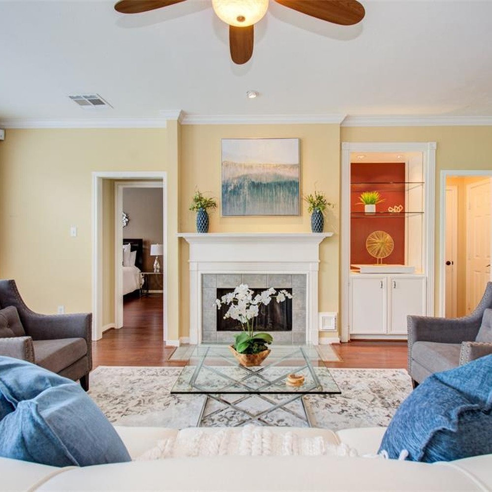 Living room with two blue pillows on white sofa. Glass coffee table with wooden vase with white orchid. Two dark gray chairs.