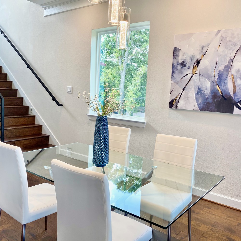 Glass dining table with four leather white chairs. Dark blue and gold artwork by window.