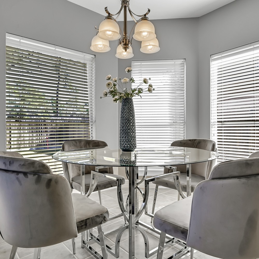 Glass breakfast table with four suede gray chairs. Blue vase with white flowers displayed.