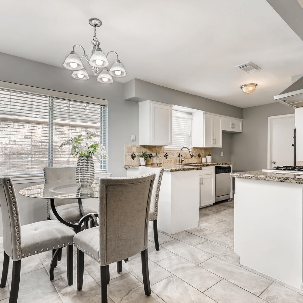 Breakfast room with glass table with black metal frame. Four gray chairs, and vase with white flowers on the table.
