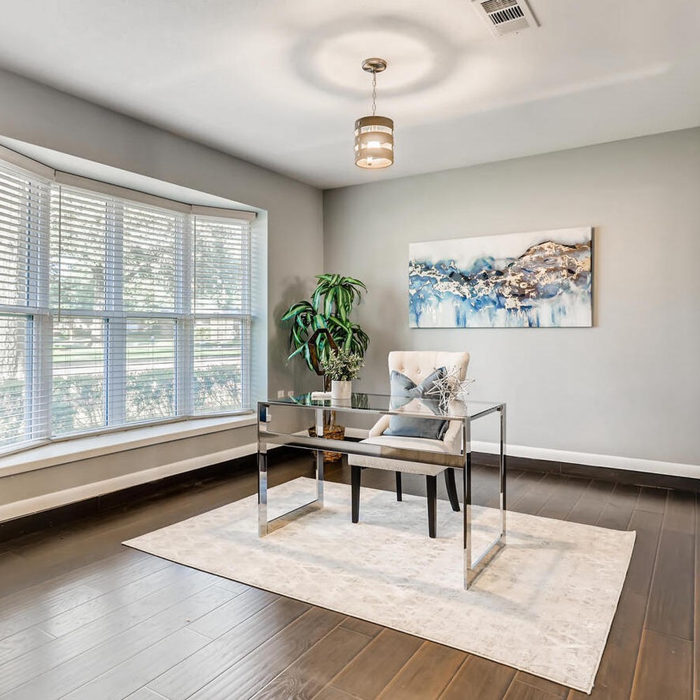Office with white rug, glass desk, and accessories. Blue and gold art behind desk, and plant in corner of room.