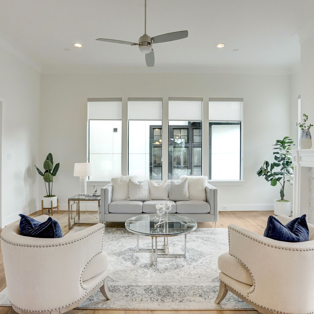 Living room with rug, sofa, glass coffee table, two plants, and two chairs looking at windows.