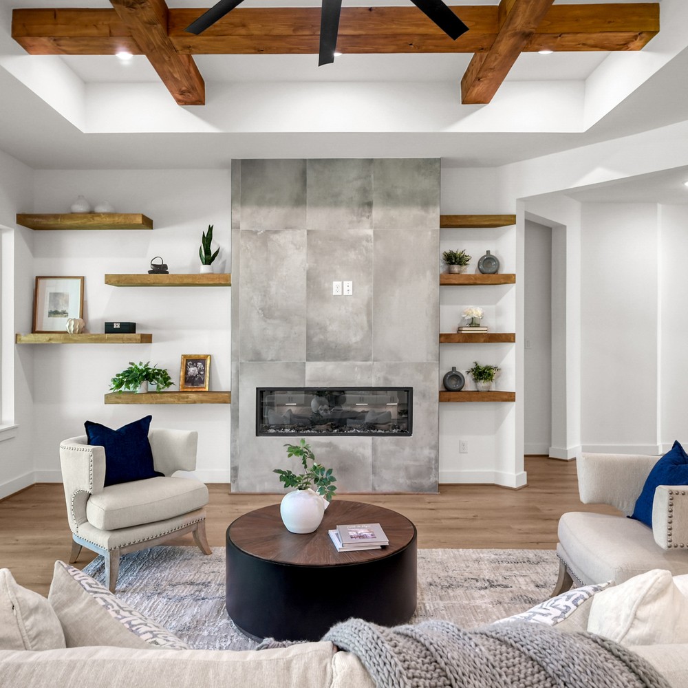 Transitional living room staged in a Conroe, TX home with wood ceiling beams, floating shelves, and a concrete-look linear fireplace.