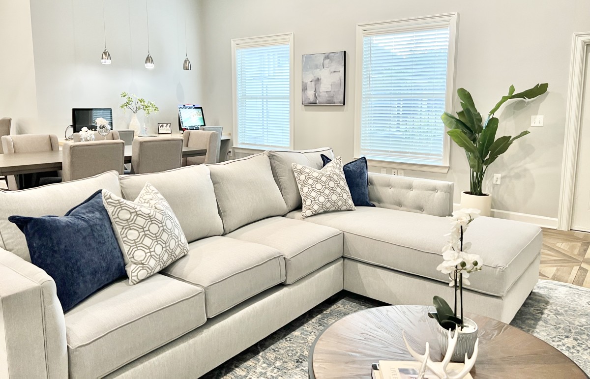 This apartment retreat features a sitting area with a sofa and coffee table. On the coffee table is an orchid and book with painted deer horns placed on top. Behind the sofa is a table with six chairs and a beautiful flower vase.