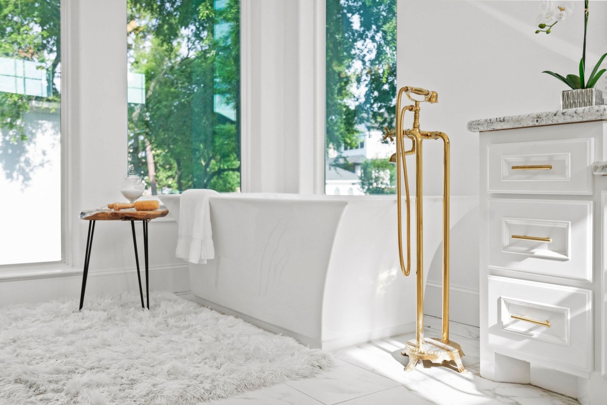 A bathroom with a white ceramic bathtub surrounded by windows. A white rug with a wooden side table is next to the tub. The water faucet is gold with a handle. The counter holds an orchid.